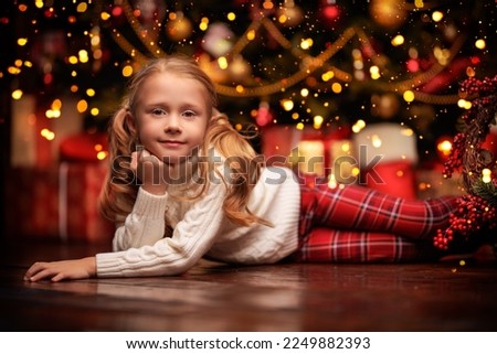 A happy little girl in a white winter sweater and red pants lying on the floor and dreamily looking at the camera. Festive Christmas interior background. New Year and Christmas dreams. 