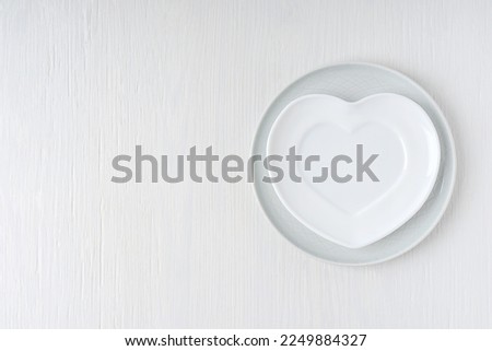 Top view of empty heart shaped plate served on round gray plate on white wooden background with copy space. Tableware preparation for celebration of valentines day or romantic dinner decoration