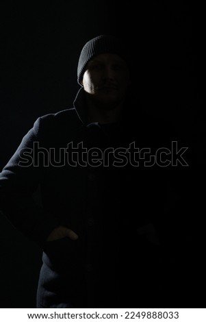 Horror concept. Studio portrait of man with dark coat and hat hiding in black shadow. Model silhouette illuminate with light. Copy space and dark studio background