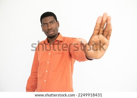 Portrait of confident young man making stop gesture over white background. African American guy wearing orange T-shirt showing forbiddance sign. Restriction concept