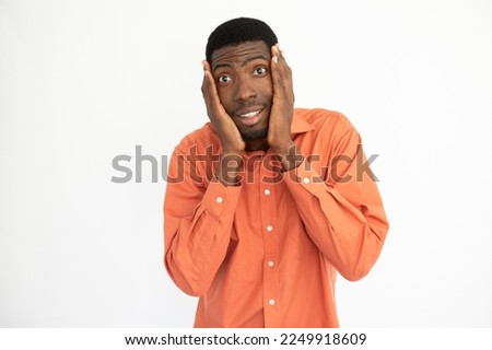Portrait of amazed young man holding face in hands over white background. African American guy wearing orange T-shirt looking at camera with awe. Surprise concept