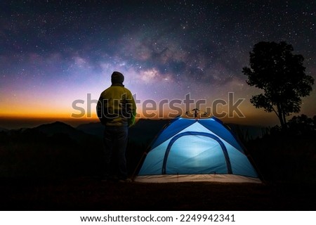 A glowing blue camping tent man standing in a high place looking up in wonder at the Milky Way galaxy
