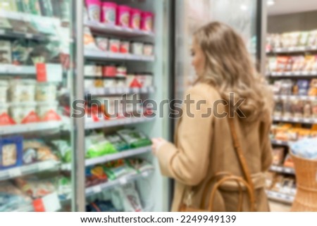 A young woman chooses dairy products in a store. A beautiful blonde in a stylish beige coat opened the refrigerator door. Healthy food and vitamins. Blurred.  Royalty-Free Stock Photo #2249949139