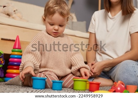 Early development. Photo of little toddler girl in beige sweater playing with educational colorful toys at home, sitting in living room near sofa with her mother.