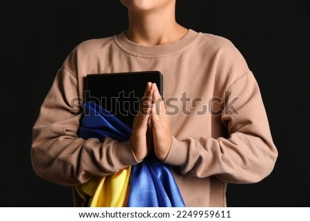 Little boy with Holy Bible and flag of Ukraine praying on black background, closeup