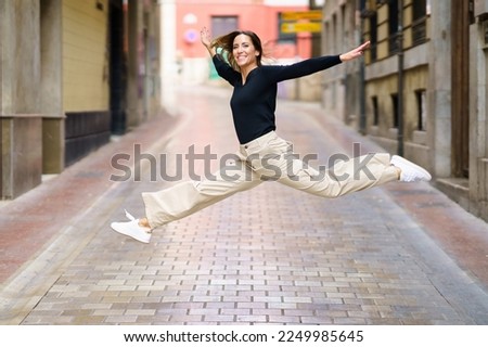 Side view of delighted young female with brown hair in casual clothes and sneakers smiling and looking at camera while jumping on narrow paved street Royalty-Free Stock Photo #2249985645