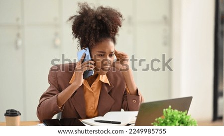 Serious African woman employee looking at laptop screen and talking on mobile phone, arguing with a client or customer