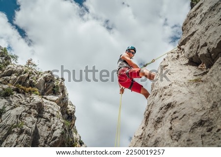 Active climber middle age man in protective helmet looking at camera while abseiling from cliff rock wall using rope with belay device and climbing harness. Active extreme sports time spending concept Royalty-Free Stock Photo #2250019257