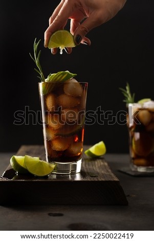 Cocktail Cuba Libre with cola, lime and ice in a glasses on a wooden board. Cola and lemon drink. Summer refreshing drink. Female hand squeezing lime into a glass of cocktail.