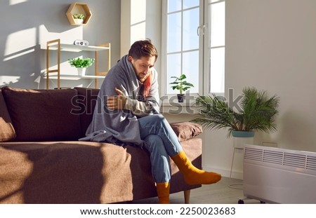 Young man covered with blanket sitting on couch beside heater trying to warm up. Shivering man suffering from cold while sitting on sofa in living room at home. No heating, power problems concept Royalty-Free Stock Photo #2250023683
