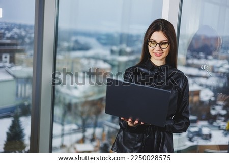 Young freelancer woman in glasses with laptop working remotely in modern workspace with large windows. Remote work