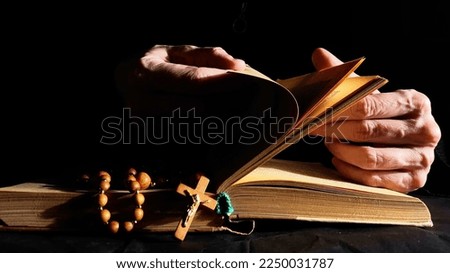 Hands flipping Bible in dark. Book, cross, rosary on black background. 