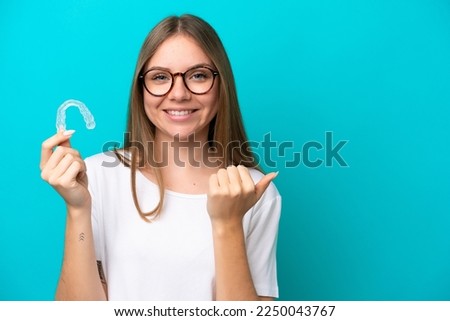 Young Lithuanian woman holding invisible braces isolated on blue background pointing to the side to present a product