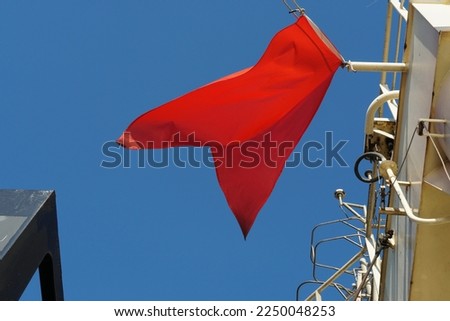 Red flag, international maritime signal flag, flying on the navigational mast of merchant vessel. This Bravo sign means, I am taking in or discharging or carrying dangerous goods. Behind is blue sky. Royalty-Free Stock Photo #2250048253