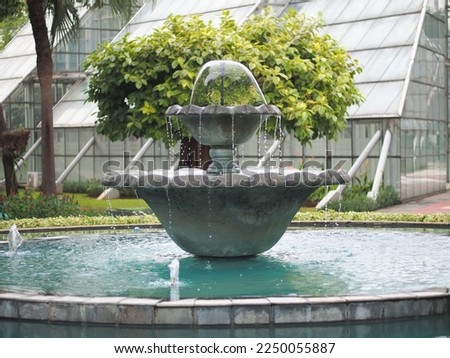 a fountain in the middle of a City park with trees and greenhouses in the background