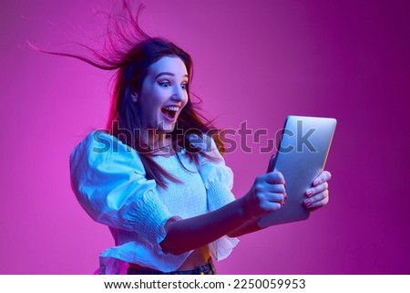 Portrait of young emotive girl cheerfully looking on tablet over pink background in neon light. Concept of youth, beauty, fashion, lifestyle, emotions, facial expression. Ad