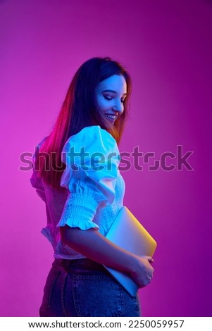 Portrait of young girl, student in white blouse posing with laptop over purple background in neon light. Online education. Concept of youth, beauty, fashion, lifestyle, emotions, facial expression. Ad