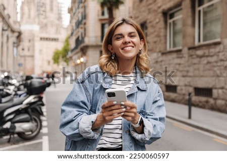 Picture of happy cute woman looking at camera with phone in hands. Smiling blonde staying on the city background. Street style. technology concept