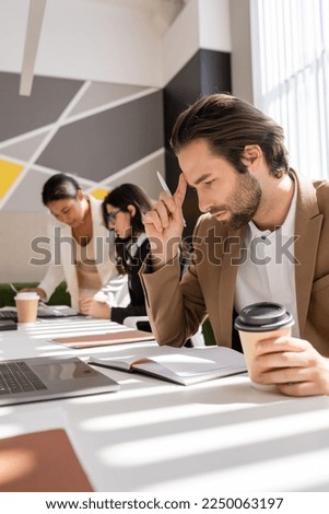 thoughtful businessman with coffee to go looking in notebook near interracial colleagues working on blurred background