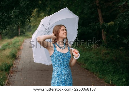 A slender young smiling girl with blond long hair, in a blue summer dress and blue beads, stands with a white umbrella in her hands, on a path in a forest park area in the rain.