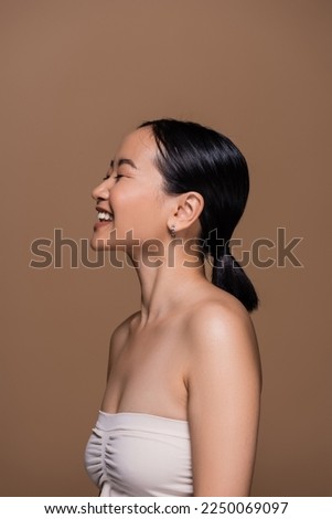 Young asian woman in top and with ponytail hairstyle standing isolated on brown