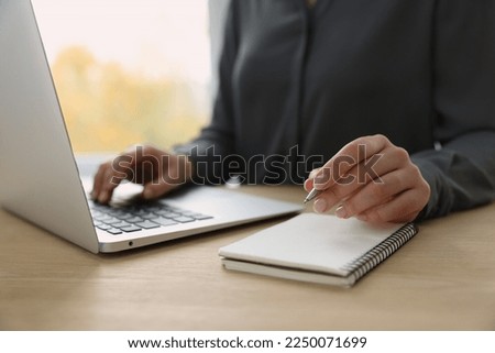 Woman with notebook and pen working on laptop at wooden table, closeup. Electronic document management