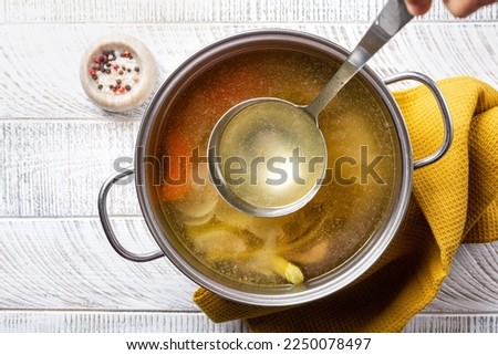 Cooking beef broth stock in a saucepan with a ladle , made using meat, bones, vegetables, carrot, onin, celery, and seasoning. 