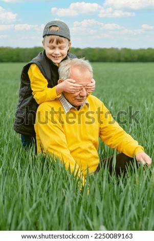 Family activity outdoors game and playtime positive. The boy closes his eyes to the grandfather sitting on the green grass. High quality photo Royalty-Free Stock Photo #2250089163