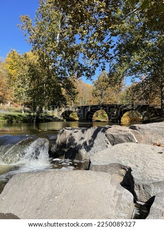 The historical Stone Arch Bridge in the Catskills. The stone footbridge has three arches, is full of falling, Autumn leaves, and green grass. A small waterfall is ahead.
