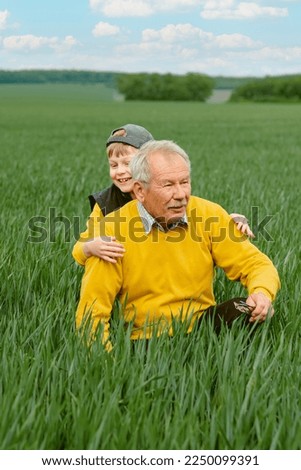 An elderly man sitting on the green grass, his grandson hugging him behind. Family activity outdoors game and playtime positive. . High quality photo Royalty-Free Stock Photo #2250099391