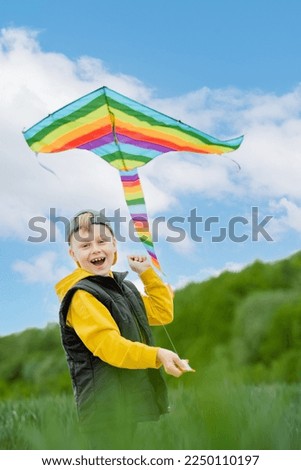 Focus on the child's hands holding a rainbow kite in the sky and looking at the camera. Active outdoors game and playtime positive. High quality photo Royalty-Free Stock Photo #2250110197