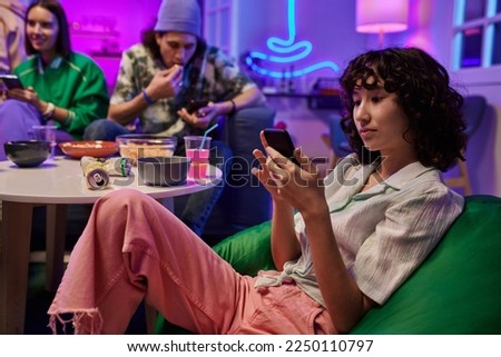 Young gorgeous brunette woman in white casual blouse and pink jeans scrolling in smartphone while relaxing in armchair against friends Royalty-Free Stock Photo #2250110797