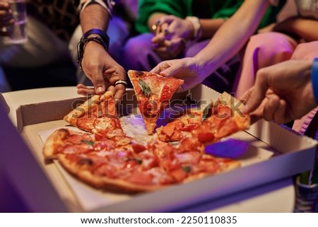 Close-up of slices of appetizing pizza in square cardboard box and hands of young friends taking them and eating while enjoying home party
