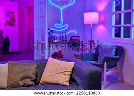 Spacious living room lit by neon light with soft comfortable couch in the center surrounded by armchair and small table with alcoholic drinks Royalty-Free Stock Photo #2250110863