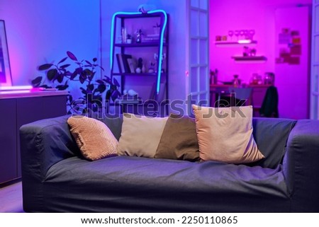 Comfortable sofa with row of soft cushions standing in the center of living room lit with neon light prepared for guests and home party Royalty-Free Stock Photo #2250110865