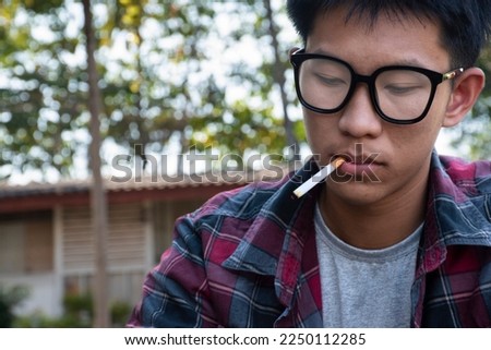 Young asian teen boy in plaid shirt wears rainbow wristband and holds cigarette in hand and smoking, blurred background, concept for bad behavior of young teens all over the world. Royalty-Free Stock Photo #2250112285