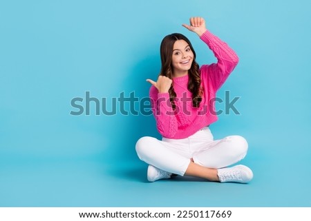 Full length photo of charming girl dressed knit pullover sitting legs crossed pointing thumbs empty space isolated blue color background