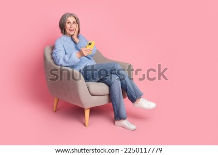 Full size photo of funny senior grandmother sit armchair blue knitted pullover touch cheek shock many followers isolated on pink color background