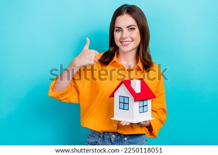 Photo of adorable confident lady wear orange shirt holding small house showing thumb up isolated turquoise color background