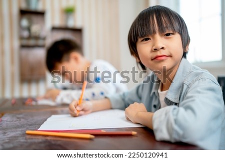 Portrait of Asian boy sit with look at camera while his friend concentrate to drawing or painting the picture in the back of classroom.