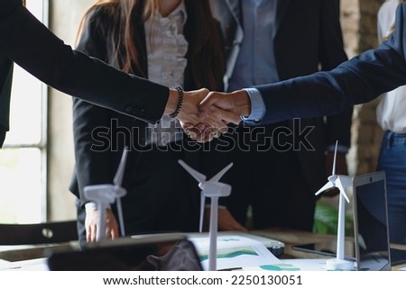 Handshake between two business partners, surrounded by wind turbine models and charts. Ideal for illustrating themes of partnership, renewable energy, and sustainability - business lifestyle Royalty-Free Stock Photo #2250130051