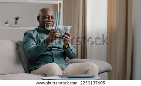 Happy old bald overweight African American senior mature elderly man happy with financial savings salary at home sitting couch. Winning online lottery holding dollars throw up cash money Royalty-Free Stock Photo #2250137605