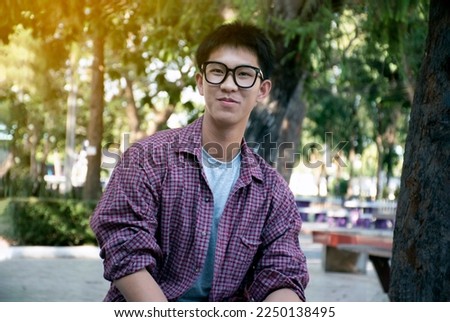 Portrait young asian handsome boy in plaid shirt wears eyeglasses, smiling and sitting on bench in rest area of high school, raising teens and easy going life of teenagers concept. Royalty-Free Stock Photo #2250138495