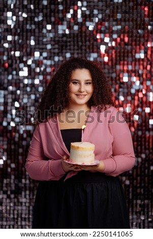 Attractive stylish plus size model with curly cool hairstyle in pink clothes is holding a cake on a shiny pink background.