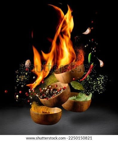 Hot spices and chili peppers in wooden bowls flying over black background and fire. Spices and seasonings powder with flame splash. Freeze motion photo. Royalty-Free Stock Photo #2250150821