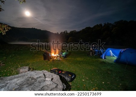 Blue Camping Tent Illuminated Inside. Night Campsite. Recreation. Motorcycle traveler, tourist bikers. Mist, moon and stars. take a rest after long riding. Equipment for travel. Campfire, bonfire