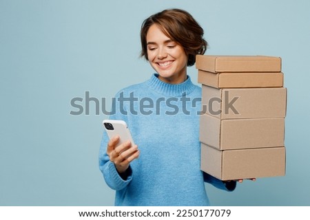 Young happy caucasian woman wear knitted sweater hold stack cardboard blank boxes use mobile cell phone isolated on plain pastel light blue cyan background studio portrait. People lifestyle concept Royalty-Free Stock Photo #2250177079