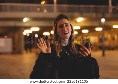 Female vlogger record with mobile phone showing hi or bye. Smiling woman taking video call on light night city. Traveler making video for her blog. Girl wave hand and look at camera, hold mobile phone