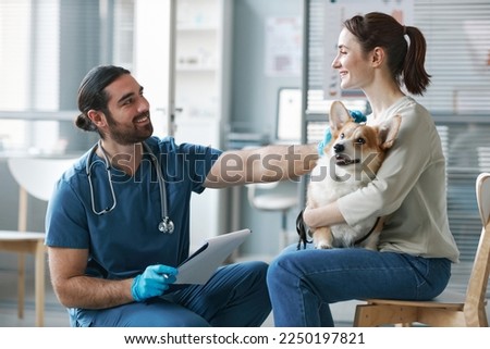 Happy young veterinarian in gloves and uniform consulting female owner of purebred welsh pembroke corgi dog in medical office Royalty-Free Stock Photo #2250197821