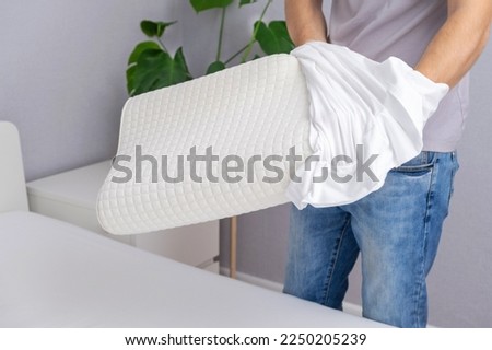 A man tucks an orthopedic pillow with a fresh bright white pillowcase. Making bed with fresh bed linen by white man. The day of change of bed linen. The day of washing bed linen in the laundry room. Royalty-Free Stock Photo #2250205239